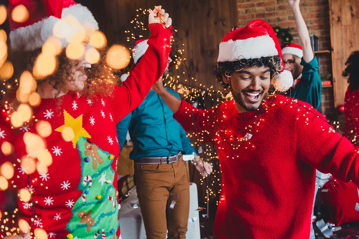 Friends in Christmas jumpers and Santa hats dancing with fairy lights, celebrating early Christmas drinks deals and festive gifting from Bargain Booze.