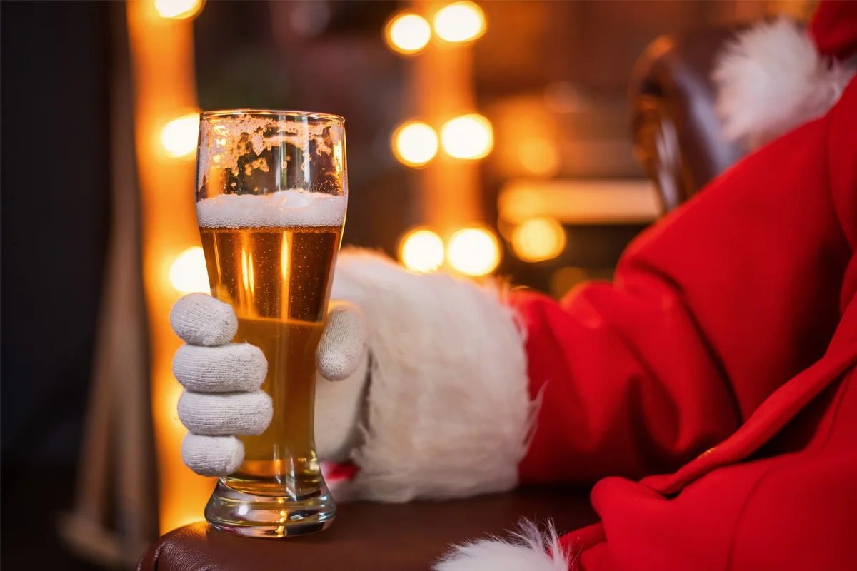 A person dressed in a Santa suit holding a pint of beer, set against warm glowing festive lights.