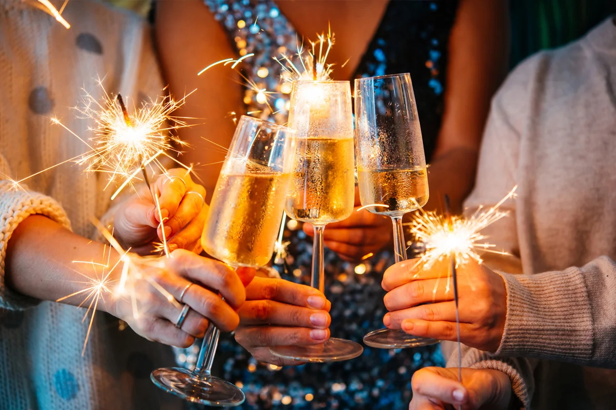 Hands holding sparklers and clinking champagne flutes at a party, with glittering outfits in the background.