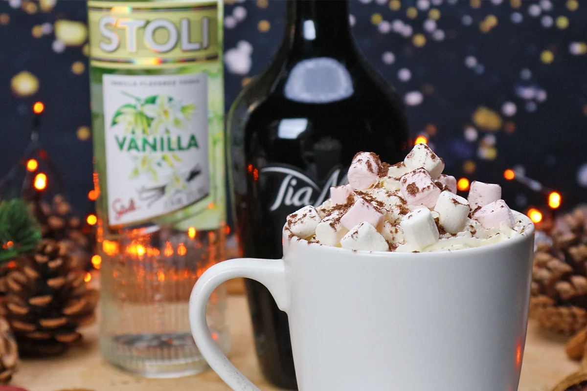 A mug of hot chocolate topped with marshmallows and cocoa powder, with bottles of vanilla vodka and coffee liqueur behind it.
