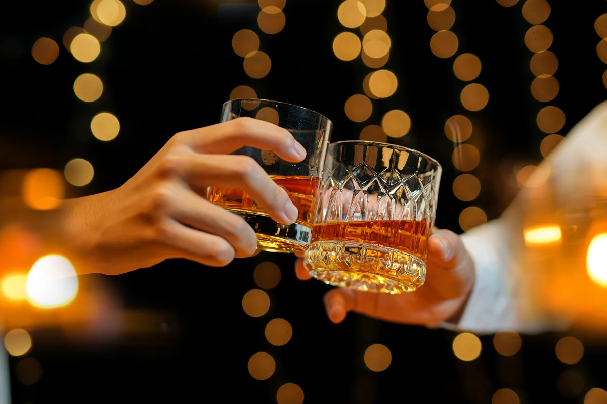 Two people clinking whisky glasses against a backdrop of golden bokeh lights, creating a festive celebratory mood.