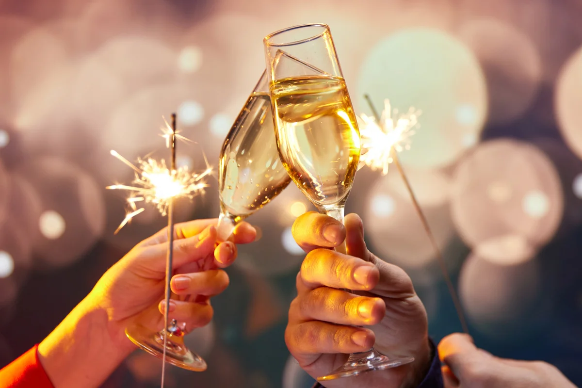 Hands holding champagne flutes and sparklers, raised in a celebratory toast against a glowing festive backdrop.