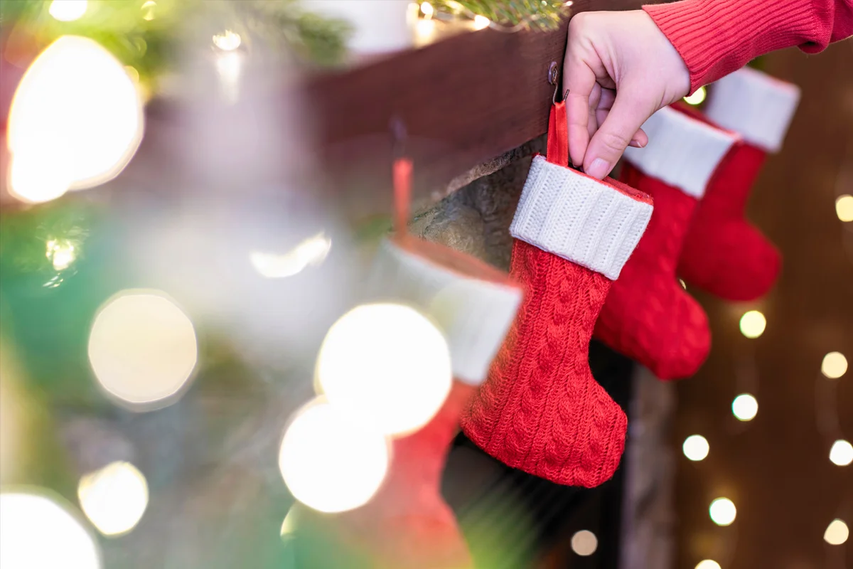 A hand placing a red knitted Christmas stocking on a fireplace mantle decorated with festive lights.
