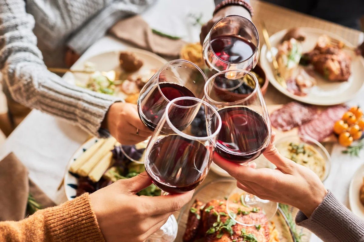 Four hands clinking glasses of red wine over a festive dinner table filled with seasonal dishes.