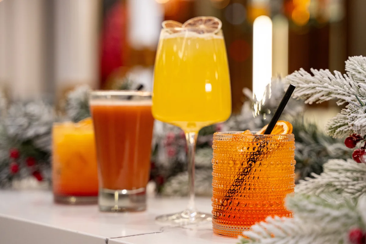 A selection of brightly coloured festive cocktails displayed on a snowy, winter-themed surface with citrus garnishes.